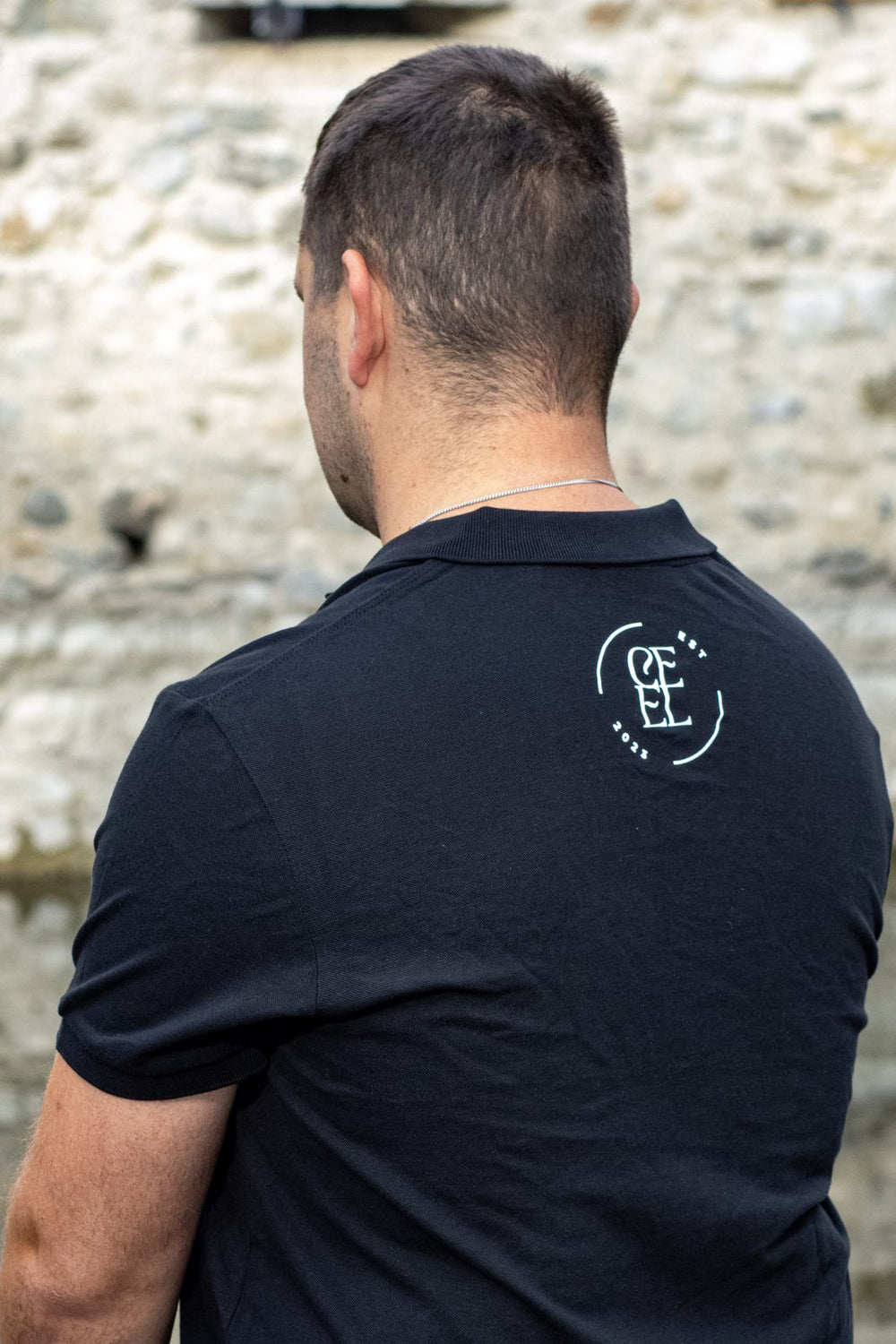 CEEL Polo Rückansicht mit Logo: Joel im schwarzen Polo von hinten, vor einer Steinmauer